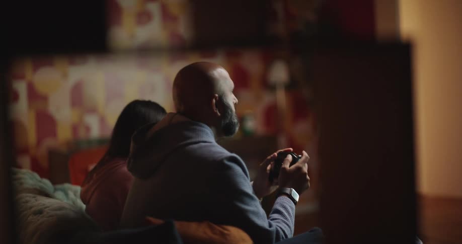 A man with a beard and a woman sit on a sofa in a dimly lit room while the man holds a game controller and looks toward a screen