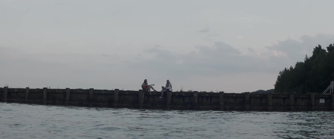 Two women sit on a dock by a body of water and one plays guitar