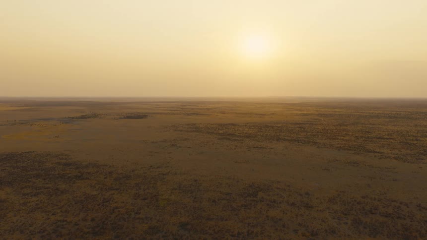 Wide aerial view of a vast desert landscape under a hazy orange sunset with the sun glowing low on the horizon