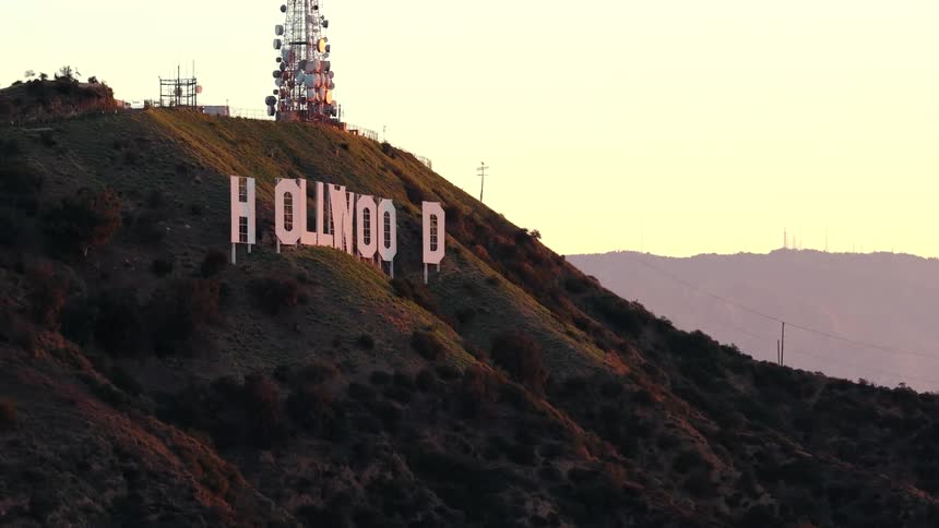 A view from the side shows the white hollywood sign on a green and brown hill with a tall radio tower and mountains in the background during sunset