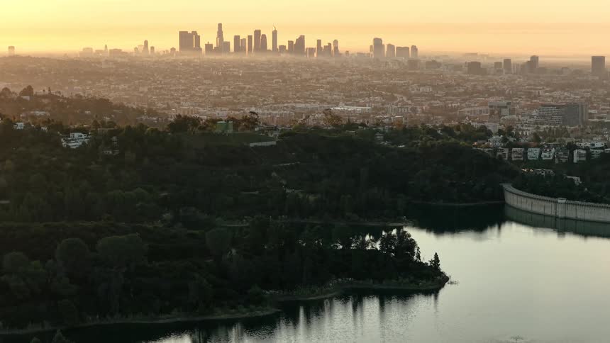 The camera pans slowly across a serene reservoir in the foreground bordered by lush greenery and a concrete dam structure in the background the hazy silhouette of a major city skyline sits under the warm golden glow of a setting sun