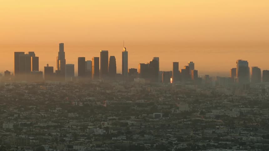 A wide aerial view captures the silhouette of a city skyline with skyscrapers rising above a hazy urban sprawl under a golden orange sunset sky