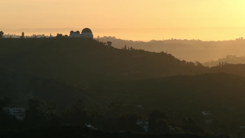 The griffith observatory sits atop a hazy hill as the sun begins to rise over the surrounding california landscape the golden morning light casts the rolling terrain into deep silhouette while a soft mist settles into the valleys below