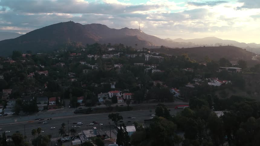 A highway filled with flowing traffic at the base of a lush hilly neighborhood while the sun sets behind distant mountain peaks under a soft clouded sky highlighting the transition between urban life and nature