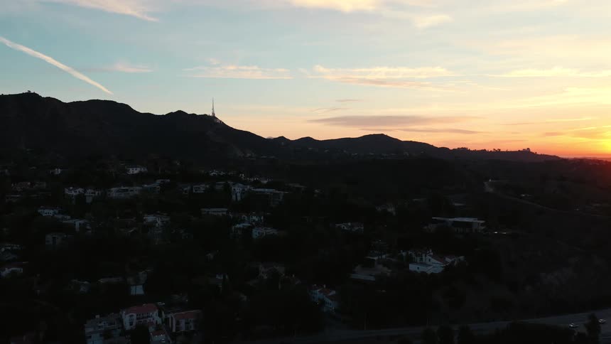 A wide aerial view shows a hillside neighborhood at dusk as the sun sets behind distant mountain silhouettes under a hazy orange and blue sky