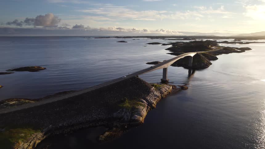 A high aerial view shows a curved concrete bridge connecting rocky islands over calm ocean water under a bright morning sky with soft sunlight