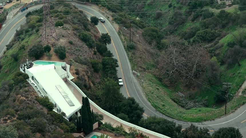 An aerial shot captures cars driving along a winding canyon road next to a modern hillside home with a swimming pool