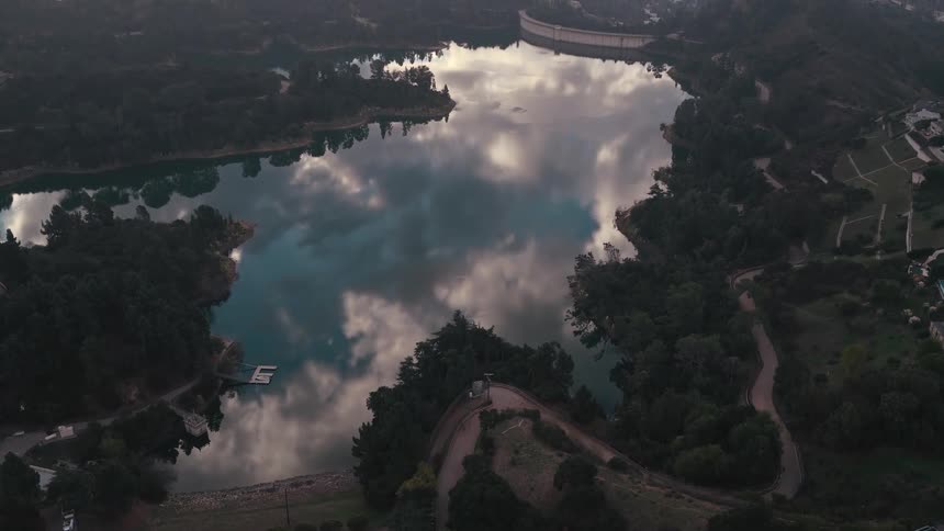 The camera provides an overhead view of a large reservoir that perfectly reflects the cloudy sky surrounded by dense trees and winding hillside roadsas the view expands a distant city skyline is revealed beneath a hazy golden sunset that casts a wa
