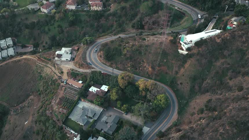An aerial topdown view shows a white vehicle navigating a sharp ushaped curve on a paved road that winds through a steep brushcovered hillside dotted with luxury homes