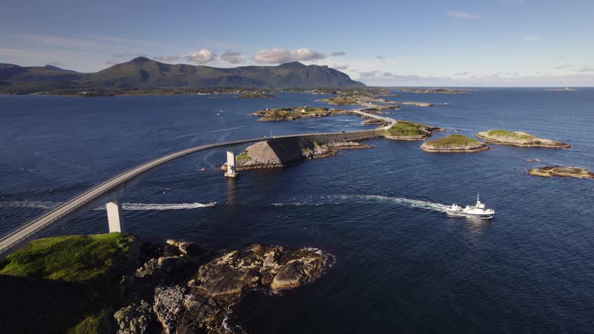 A boat glides under a bridge that connects a series of small islands crossing the bright ocean at midday with sunlight reflecting off the water
