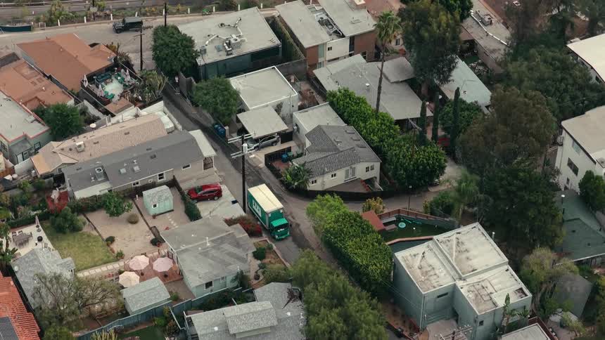Green delivery truck drives along a narrow residential street lined with various suburban homes and lush green treeshe camera pans upward to reveal a heavily congested multilane highway running parallel to the quiet neighborhood