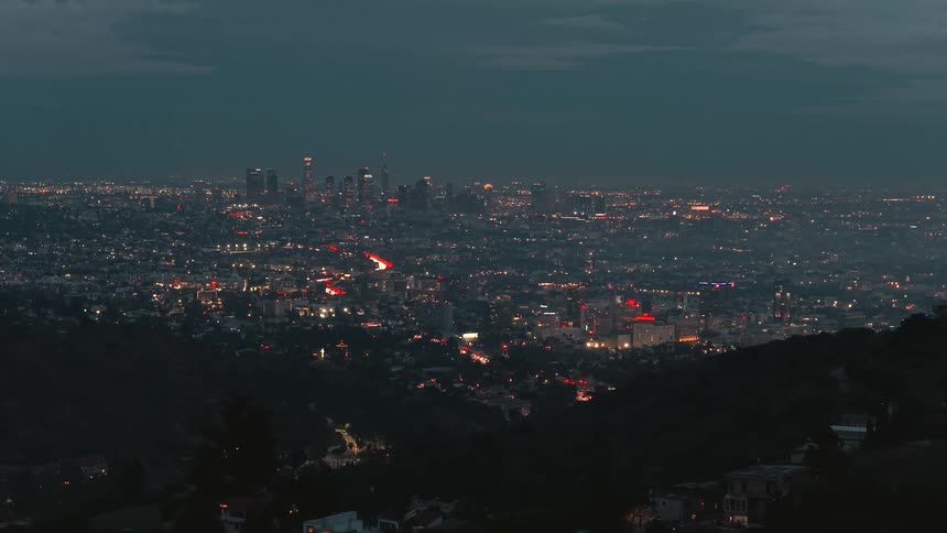 A highangle long shot overlooks a vast city basin at night revealing a dense carpet of glowing city lights and red taillight streaks from traffic that lead toward the prominent illuminated skyscraper cluster of the downtown skyline