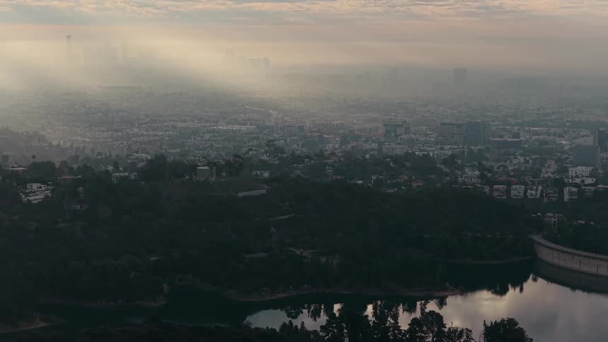 Sunlight streams through a thick layer of haze over a vast city nestled among rolling treecovered hillsin the foreground a calm body of water reflects the surrounding dark greenery and the soft morning sky