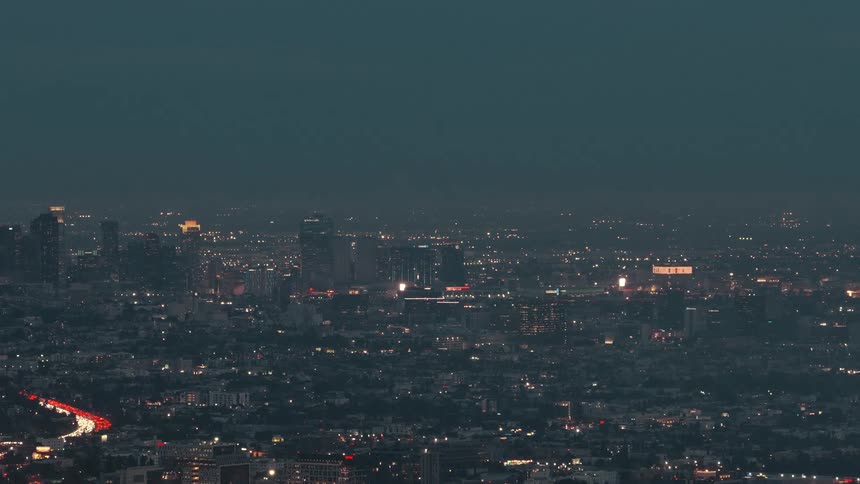 From a high vantage point a vast cityscape twinkles with countless lights as a vibrant stream of traffic flows through the urban expanse under a dark sky