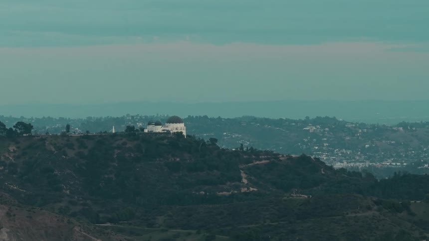 The white facade and dark domes of an observatory perched atop a rugged brushcovered hill overlooking the sprawling urban basin of a city under a pale overcast sky
