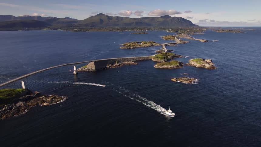 An incredible aerial view shows a long winding bridge connecting a series of rocky islands as boats cruise across the deep blue water with majestic mountains on the horizon