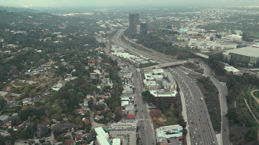 An expansive aerial view captures a major freeway slicing through a vast urban landscape with cars flowing past hillside homes and towering city buildings in the distance