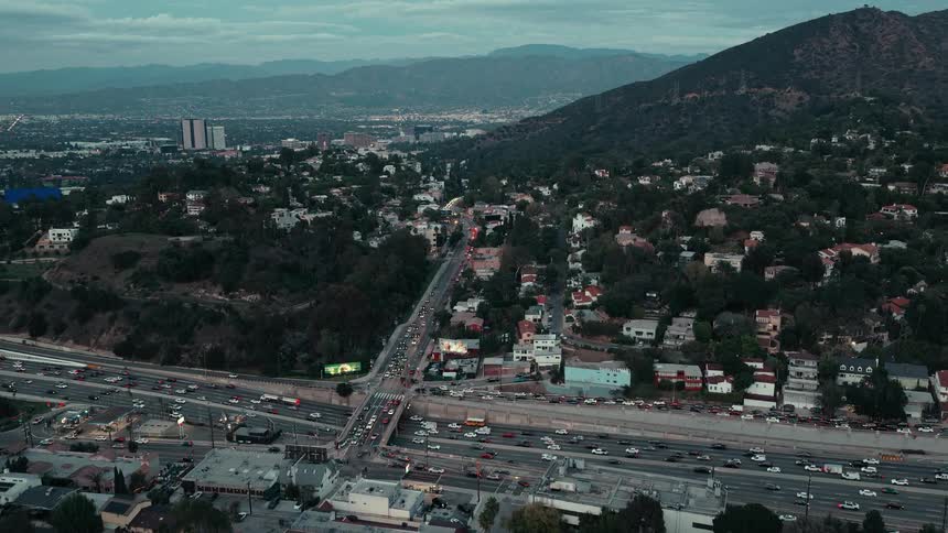 An aerial shot captures a busy highway intersection nestled between a residential hillside and a vast valley under a cloudy dusk sky lines of traffic move slowly through the urban landscape as the city lights begin to flicker on in the distance
