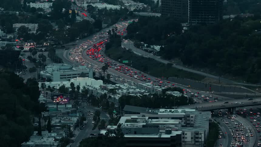 A highangle view captures heavy evening traffic on a winding city highway where red and white vehicle lights contrast sharply with the darkening urban landscape and surrounding buildings during twilight