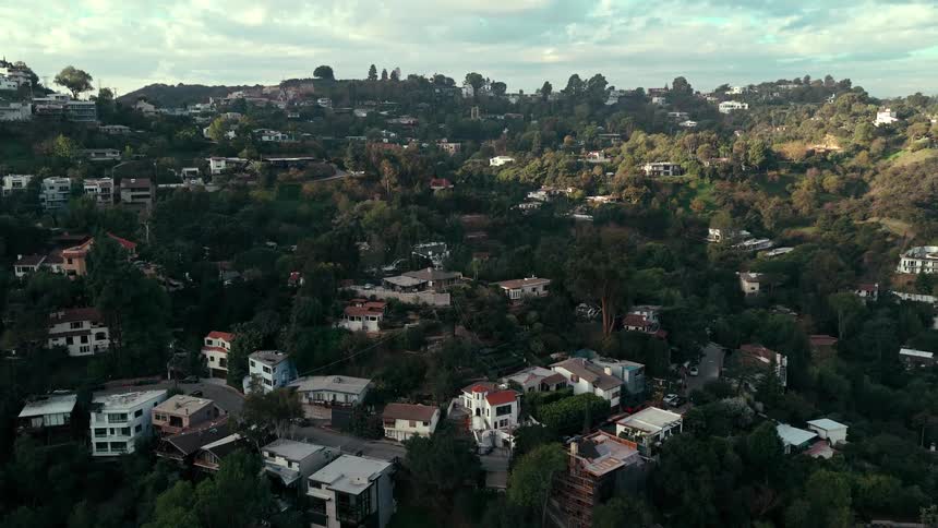 An aerial tracking shot glides over a steep verdant hillside populated with various modern and traditional houses nestled among thick trees and winding roads during the soft lighting of late afternoon
