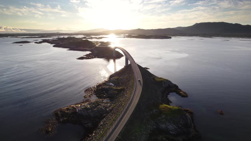 Cars travel on a road with a bridge connecting islands as the sun shines brightly over mountains along the coast