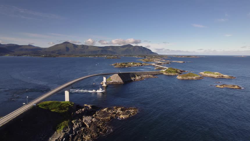 An aerial view shows a white boat passing under a bridge which winds across small rocky islands surrounded by blue water against a backdrop of distant coastal mountains