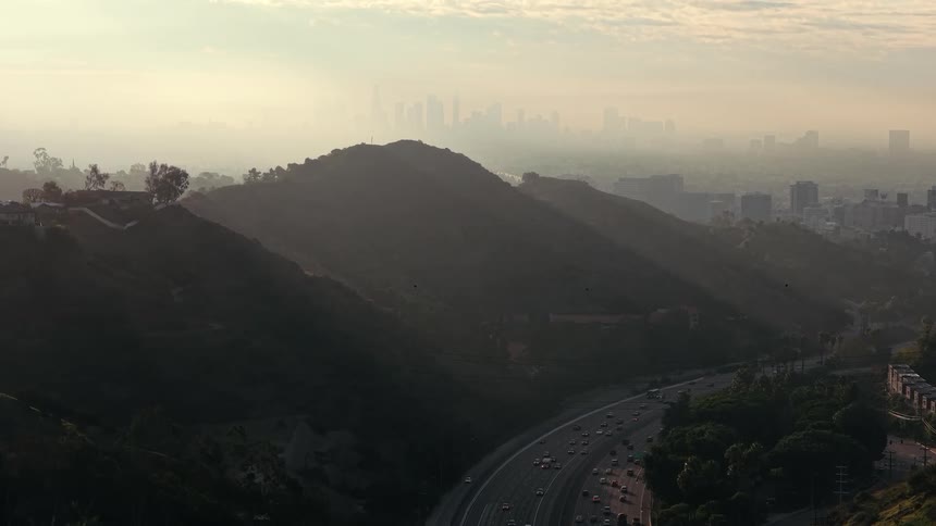 In this scene a multilane highway curves along the base of a dark shadowed hillside during the early morning hours in the background the distant city skyline is partially obscured by a thick layer of haze and soft golden light