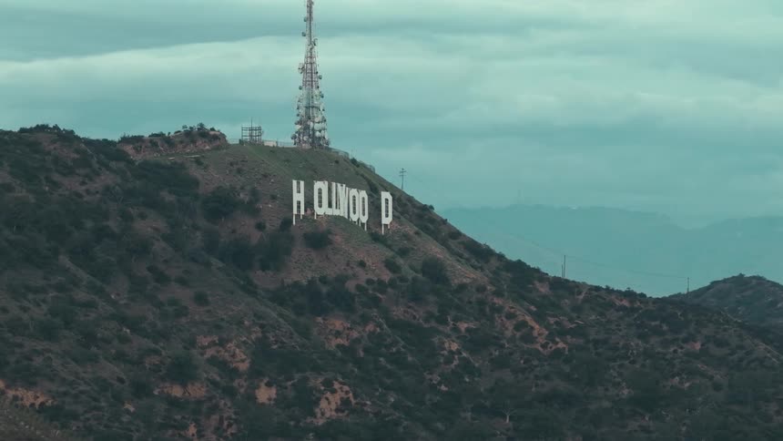 Hollywood sign mounted on a hillside with a transmission tower in the background under a cloudy sky