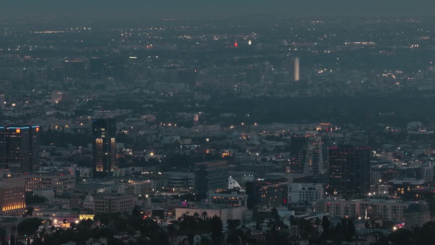 A highangle view captures a sprawling metropolitan landscape at twilight showing a dense collection of illuminated skyscrapers apartment buildings and city lights extending toward a distant white monument under a dark hazy evening sky