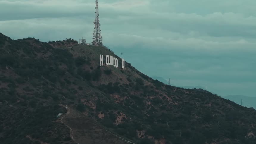 This aerial shot captures the iconic hollywood sign nestled on the rugged brushcovered slopes of mount lee under an overcast sky a tall radio tower stands prominently on the peak above the white capital letters as the camera slowly pans across the hill