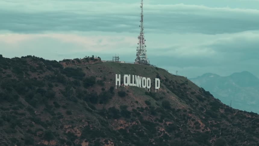 The hollywood sign is situated on a steep rugged hillside with a tall communications tower positioned directly behind it the surrounding landscape is characterized by rolling scrubcovered mountains under a hazy overcast sky