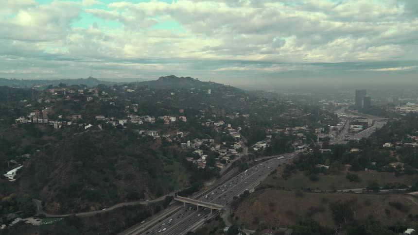 In this aerial video two lines of heavy traffic flow in opposite directions along a wide multilane highway the highway cuts through a hilly residential landscape under a vast cloudfilled sky