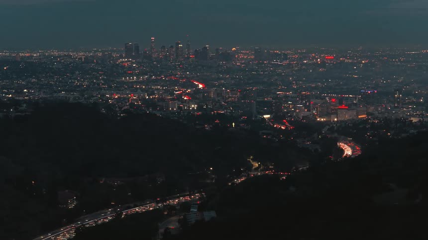 An aerial view captures the sprawling los angeles cityscape at night with streams of traffic lights flowing through the dark hills and the downtown skyline visible in the distance
