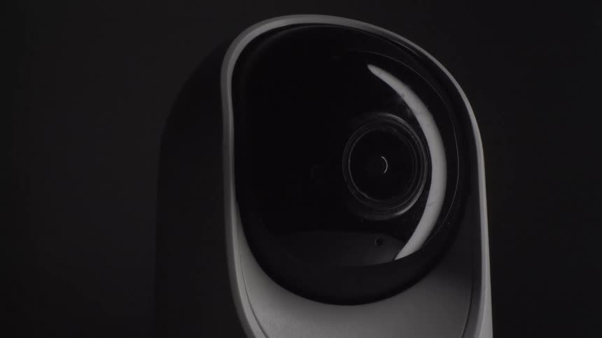 A white indoor security camera sits in a dark environment with its black spherical lens housing facing forward the camera lens slowly pans from its right toward the center against the shadowed background