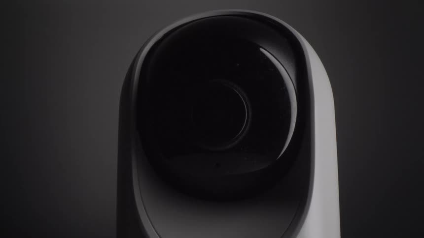 A white indoor security camera with a large black circular lens slowly rotates its head against a dark minimalist background to monitor the surrounding environment