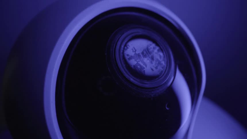 This video features a closeup cinematic shot of a modern security camera rotating slowly under deep blue lighting the camera lens moves toward the viewer reflecting light across its glass surface as it pans into a direct frontfacing position