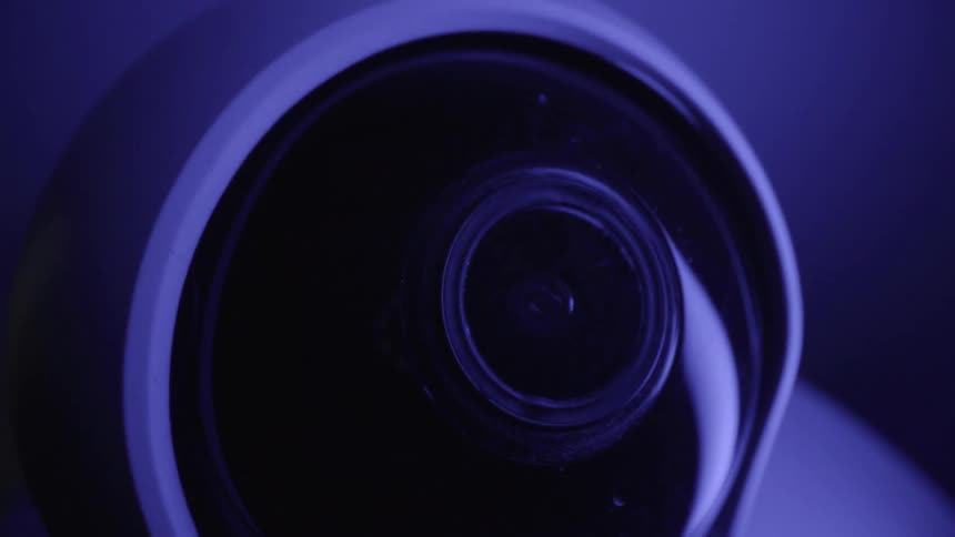 A closeup shot of a white dome security camera as its internal lens rotates and tilts upward under blue ambient lighting to monitor its surroundings