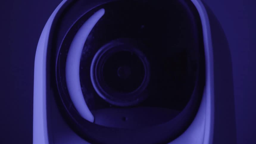Bathed in cool blue light a white smart security camera rotates its black lens housing horizontally and vertically