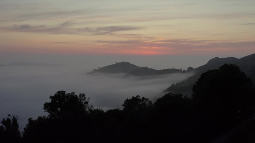 Silhouetted trees and some people overlook a vast valley filled with thick white fog during a quiet sunrise as the camera slowly pans right to reveal rolling mountain ridges emerging from the soft morning mist