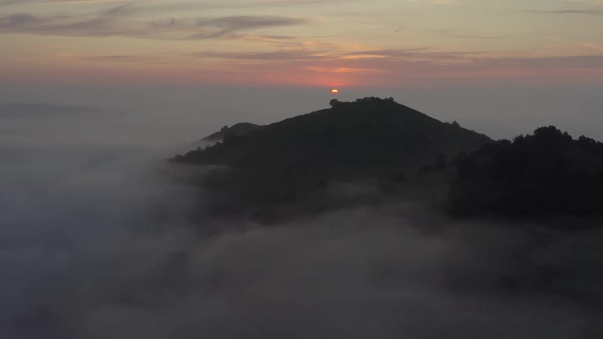 Thick fog fills the forested mountain valley as the sun shines through clouds on the horizon