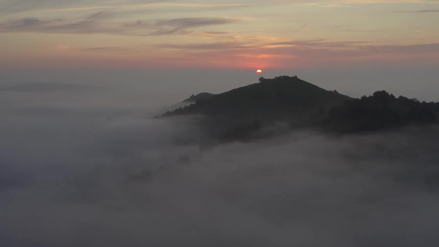 Partially visible sun over treecovered hill shrouded in mist at dawn