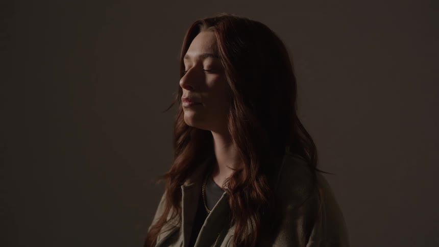 A young woman wearing a green jacket stands with her eyes closed deep in thought