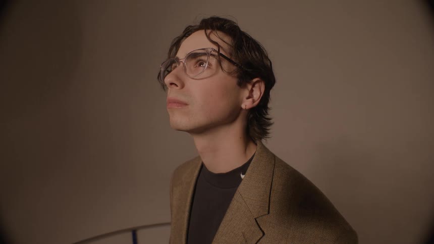In a dimly lit warmtoned room a young man with glasses and a tweed jacket looks up with a pensive expression as light moves across his face