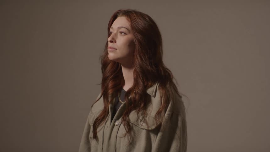 A young woman stands indoors with long hair wearing a green jacket as a burst of bright light briefly illuminates the room