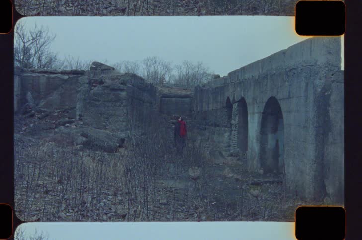 A young woman wearing a jacket stands inside a ruined building at midday during the fall season surrounded by scattered debris and autumn light
