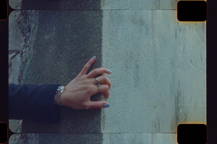 A hand with rings and blue nail polish reaches out and touches a vertical seam on a textured grey wallthe footage is presented in a vintage film style featuring visible sprockets grain and color distortions that flicker across the screen