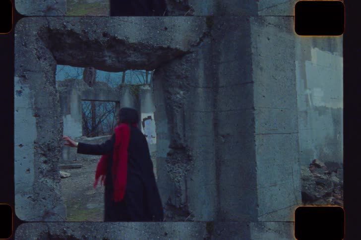 A young woman wearing a black coat and a red scarf walks into a roofless ruined building looking around as she explores the space