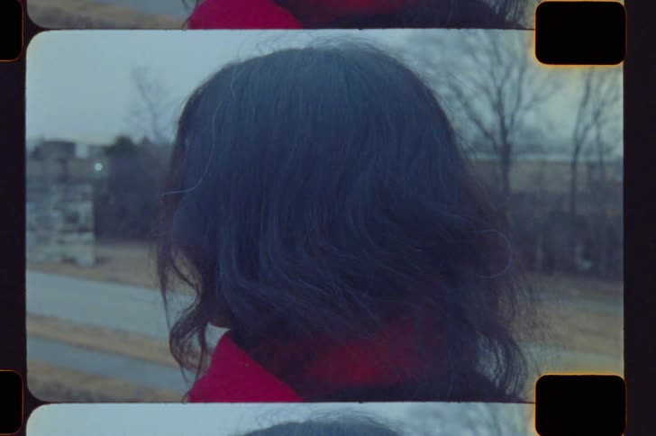 A woman with long dark hair and a red scarf stands outside on a cold day looking away from the camera before slowly turning her head to look back toward the left in a profile view against a wintery landscape with bare trees