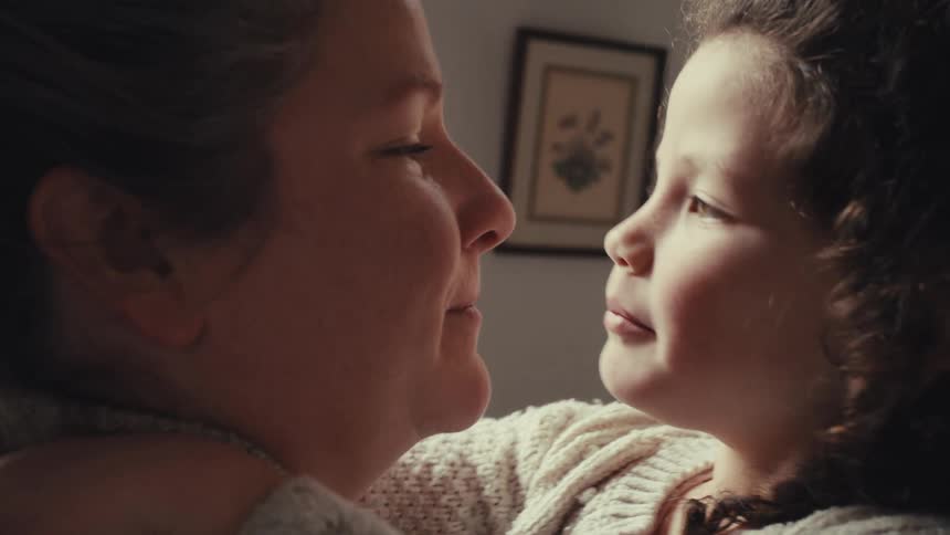 Adult woman holding and kissing a small female child who kisses her back and smiles indoors during day time