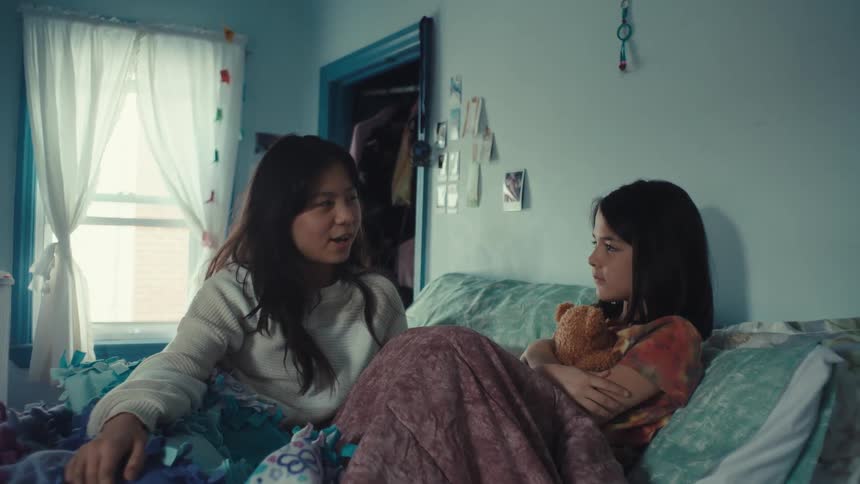 In a cozy blue bedroom a young woman sits on the edge of a bed and speaks warmly to a young girl the girl listens intently while hugging a teddy bear eventually breaking into a bright smile as they share a quiet moment